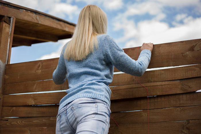 Woman in a blue sweater leaning over a wooden fence, representing Karen petty fence revenge concept outdoors.
